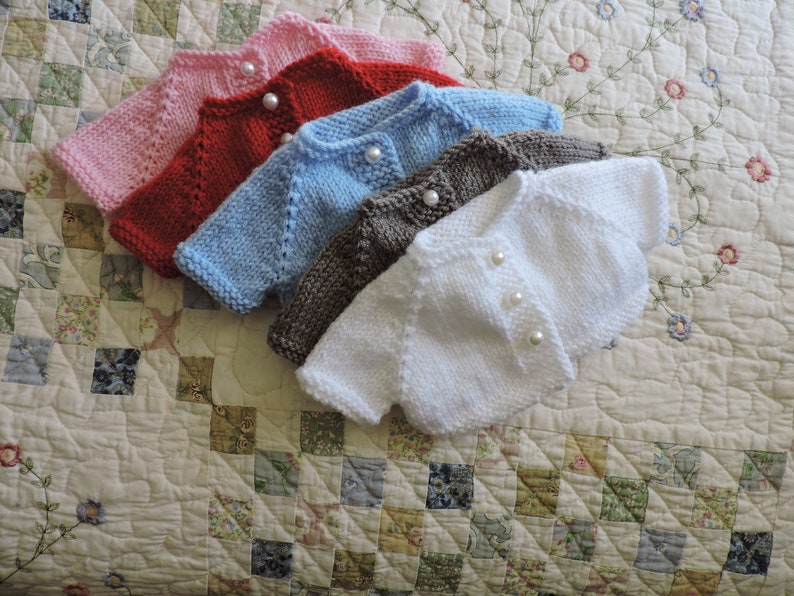 May include: Five knitted baby cardigans in different colors: pink, red, light blue, brown, and white. Each cardigan has pearl buttons.
