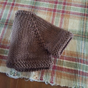 May include: A brown knitted hat with a textured pattern. The hat is lying on a plaid fabric.
