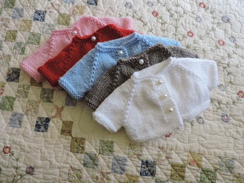 May include: Five knitted baby cardigans in different colors: pink, red, light blue, brown, and white. Each cardigan has pearl buttons and a simple design.