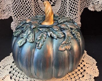 May include: A teal blue and copper pumpkin with a gold stem and leaves. The pumpkin is sitting on a white crocheted doily.