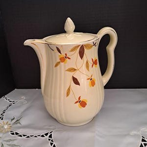 May include: Cream-colored vintage ceramic pitcher with a lid, adorned with a floral pattern in shades of brown, orange, and yellow. The pitcher has a ribbed design and a gold trim. A classic piece for serving beverages.