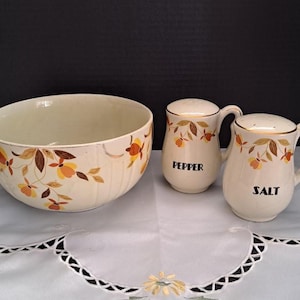 May include: A vintage cream-colored ceramic bowl and matching salt and pepper shakers. The bowl and shakers feature a floral design in shades of brown, orange, and yellow. The salt and pepper shakers have the words "Pepper" and "Salt" in black.
