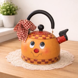 May include: A whimsical orange kettle with a smiling face design. The kettle features a black handle and spout, a red and white checkered bow, and a red and white checkered pattern around the base. It sits on a white lace doily.