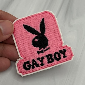 May include: Pink embroidered patch featuring a black bunny logo and the words "GAY BOY" in black. The patch has a white border and a rectangular shape with a curved bottom.