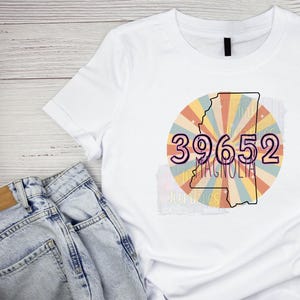 May include: White t-shirt with a retro-style graphic of the state of Mississippi with the area code 39652 and the word "Magnolia" printed on it.