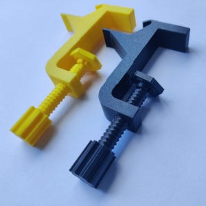 Könnte beinhalten: Zwei C-Klemmen, eine gelbe und eine schwarze, werden vor einem weißen Hintergrund gezeigt. Die Klemmen haben einen Schraubmechanismus zum Festziehen. Die gelbe Klemme befindet sich links, die schwarze Klemme rechts.