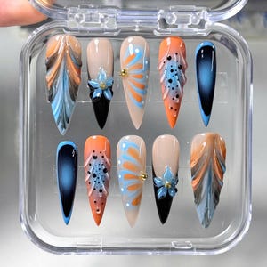 Könnte beinhalten: Ein Set aus zehn künstlichen Fingernägeln in einer transparenten Kunststoffbox. Die Nägel weisen verschiedene Designs auf, darunter abstrakte Muster in Orange, Blau und Braun, florale Akzente und Farbverläufe. Einige Nägel haben eine schwarze Basis, andere sind hautfarben.