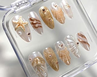 May include: A set of ten white and gold press-on nails with a beach theme. The nails feature seashells, starfish, and gold glitter.