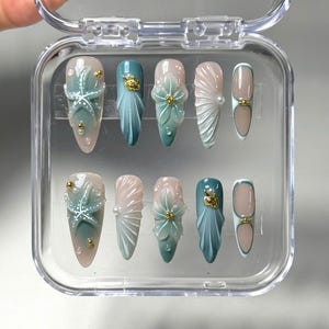 以下が含まれることがあります： 透明なプラスチックケースに入った10本のつけ爪のセット。ピンク、ティール、ゴールドの色合いで、ヒトデ、花柄、抽象的な形など、さまざまなデザインが施されています。一部の爪には、パールとゴールドのアクセントが付いています。