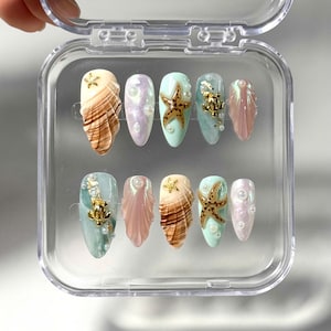 May include: A set of ten artificial nails in a clear plastic case. The nails feature various designs, including brown and white shell patterns, iridescent pink and purple, and light blue with starfish and pearl accents. Some nails have gold embellishments.