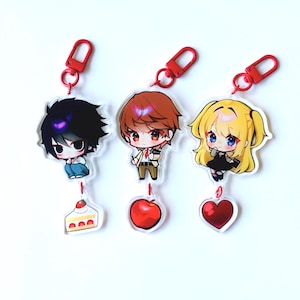 May include: Three anime-style acrylic keychains with red clasps. Each keychain features a different character with a unique charm: a slice of cake, a red apple, and a red heart. The characters have black, brown, and blonde hair.