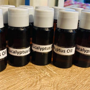 Könnte beinhalten: Eine Ansammlung kleiner, brauner Glasflaschen mit weißen Etiketten, auf denen "Eukalyptusöl" steht. Die Flaschen haben weiße Deckel und sind mit einer dunklen Flüssigkeit gefüllt. Sie stehen auf einer hellbraunen Oberfläche.