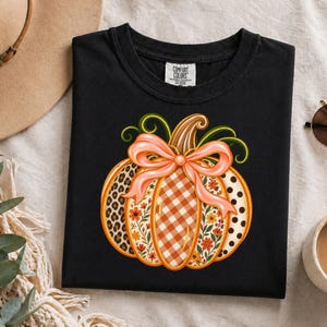 Peut inclure: Un t-shirt noir avec un motif de citrouille orné d'un nœud pêche. La citrouille présente des sections à imprimé léopard, vichy orange, motifs floraux et pois. Le t-shirt porte une étiquette Comfort Colors à l'intérieur du col.