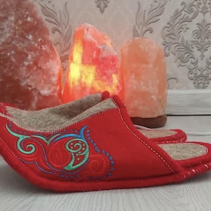 Puede incluir: Zapatillas rojas de fieltro con diseño floral bordado en azul y verde. Las zapatillas tienen un interior beige y se exhiben con tres lámparas de sal rosas iluminadas en el fondo. Un calzado acogedor.
