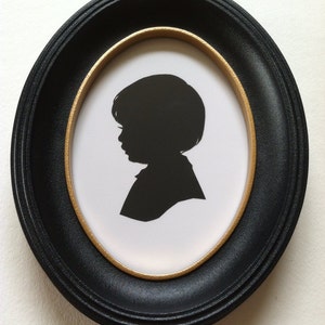 May include: A black silhouette of a child in profile, set against a white background within an oval frame. The frame is black with a gold inner border. The child's hair is short and the image is a classic portrait style.