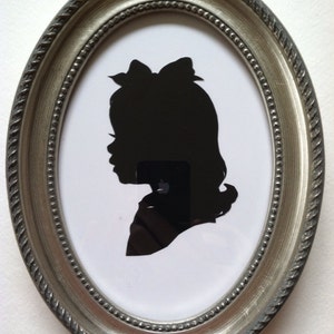 May include: Oval silver-toned frame with a black silhouette of a young person with a bow in their hair. The silhouette is set against a white background. The frame has a rope-like texture around the edge.