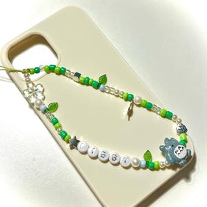 May include: A beaded phone charm with green, white, and clear beads, a star, and a gray Totoro figure. The charm spells out "L♡BBY" with white letter beads. The charm is attached to a cream-colored phone case.