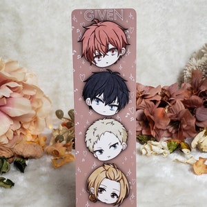 Può includere: Un segnalibro rosa e bianco con quattro teste di personaggi dei cartoni animati. I personaggi sono della serie manga "Given". Il segnalibro ha il testo "Given" in alto e "@drawsoulsmoon_" in basso.