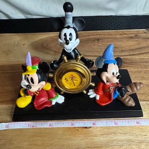 May include: A black decorative clock featuring Mickey Mouse characters. The clock has a gold-colored ship's wheel design with a yellow clock face. Mickey Mouse figures are dressed in various outfits. The base is approximately 6 inches long.