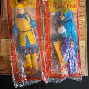 May include: Two sealed plastic packages containing action figures from Disney's "The Hunchback of Notre Dame." One figure is dressed in yellow and blue, the other in purple and blue. The packaging has red and yellow accents.