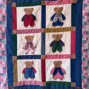 May include: A handmade quilt featuring six teddy bear appliqués, each with unique outfits in red, blue, green, and pink. The bears are set against a cream background, framed by a blue border and a patchwork design in pink and blue.