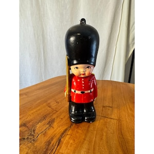 Puede incluir: Figura vintage de un guardia real británico, con un sombrero de piel de oso negro y una túnica roja con pantalones negros. La figura sostiene un rifle dorado. La cara tiene una expresión pintada.