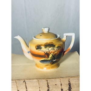Op de afbeelding: Kleine, vintage keramische theepot met deksel. De theepot is beschilderd met een landschap met een windmolen, bomen en een waterpartij, tegen een gele en oranje achtergrond. Het handvat en de tuit zijn wit.