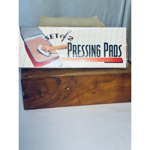 Könnte beinhalten: Eine Schachtel mit zwei Bügelkissen. Die Schachtel ist weiß mit gelben Streifen und zeigt eine Illustration einer Hand, die auf einem Kissen bügelt. Der Text "SET of 2 PRESSING PADS" ist auf die Schachtel gedruckt. Die Schachtel steht auf einer Holzoberfläche.