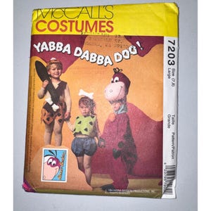 May include: McCall's Costumes sewing pattern 7203 for children, featuring Yabba Dabba Doo! text. The envelope displays three children in Flintstones-themed costumes: a cave-person outfit with a bone, a Pebbles-style set, and a purple dinosaur suit.