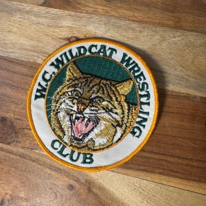 May include: A circular embroidered patch featuring a brown and tan bobcat head with an open mouth showing teeth and a pink tongue. The center has a dark green background, surrounded by a white border with the text W.C. WILDCAT WRESTLING CLUB.