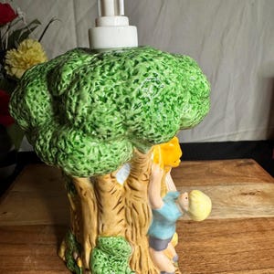 May include: A ceramic soap dispenser shaped like a tree with a green, textured top and a brown trunk. A child figure is climbing the tree. The dispenser has a white pump and is set on a wooden surface.