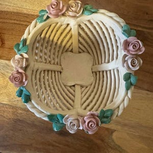 May include: Cream-colored ceramic basket with a woven design. The basket is adorned with pink and cream-colored roses and green leaves around the rim. The basket is sitting on a wooden surface.