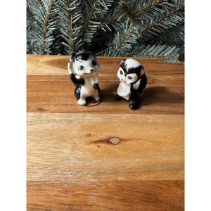 May include: Two small ceramic skunk figurines featuring black and white coloring with painted facial details. The skunks are depicted in standing positions with glossy finishes, showcasing distinct black stripes and white accents on their bodies.