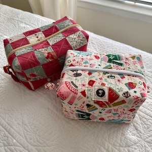 May include: Two quilted fabric pouches. One is red and green with a patchwork design and a gold zipper. The other is white with a holiday-themed pattern of mittens, hats, and mugs, and a white zipper. Both have small handles.