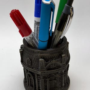 Könnte beinhalten: Ein dekorativer, dunkelbronze Stifthalter in Form eines Gebäudes, gefüllt mit verschiedenen Stiften und Markern. Die Stifte sind rot, blau, grün und schwarz. Der Stifthalter hat architektonische Details.