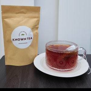 May include: A brown paper bag with the text "Today Let It Be Known" and "Known Tea Spiced Tea Blend" in a white circle. A glass teacup with a red tea inside and a tea bag string hanging out of the cup. The teacup is on a white saucer.
