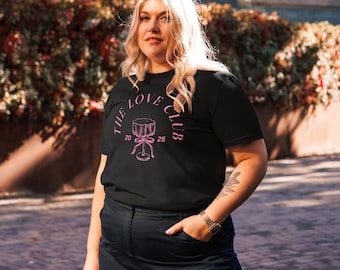 Könnte beinhalten: Schwarzes T-Shirt mit einem rosa Aufdruck, der "THE LOVE CLUB" um eine Zeichnung eines Cocktailglases mit einer Schleife zeigt. Die Zahlen "20" und "25" befinden sich auf beiden Seiten des Glases. Die Person trägt eine dunkelblaue Jeans und eine goldene Uhr.