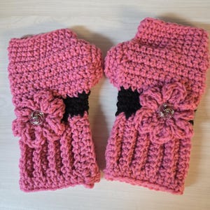May include: A pair of pink crocheted fingerless gloves. Each glove features a black band and a decorative flower with a clear button. The gloves are made with a textured stitch and are designed to keep hands warm.