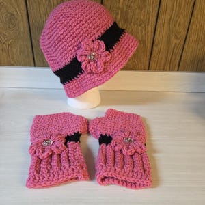 May include: A pink crochet hat and matching fingerless gloves set. The hat features a black band and a pink flower with a rhinestone center. The gloves also have black accents and flower embellishments. The set is displayed on a white surface.