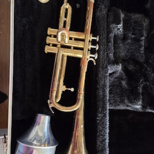 May include: A brass trumpet with a silver bell and mouthpiece, resting in an open black velvet-lined case. The trumpet has a curved design with valves and slides. The case is silver with a black interior.