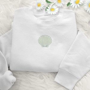 May include: A white sweatshirt with a light green embroidered seashell design on the front. The long-sleeved top is folded and placed on a white, fluffy surface. The seashell is a detailed, stylized illustration.