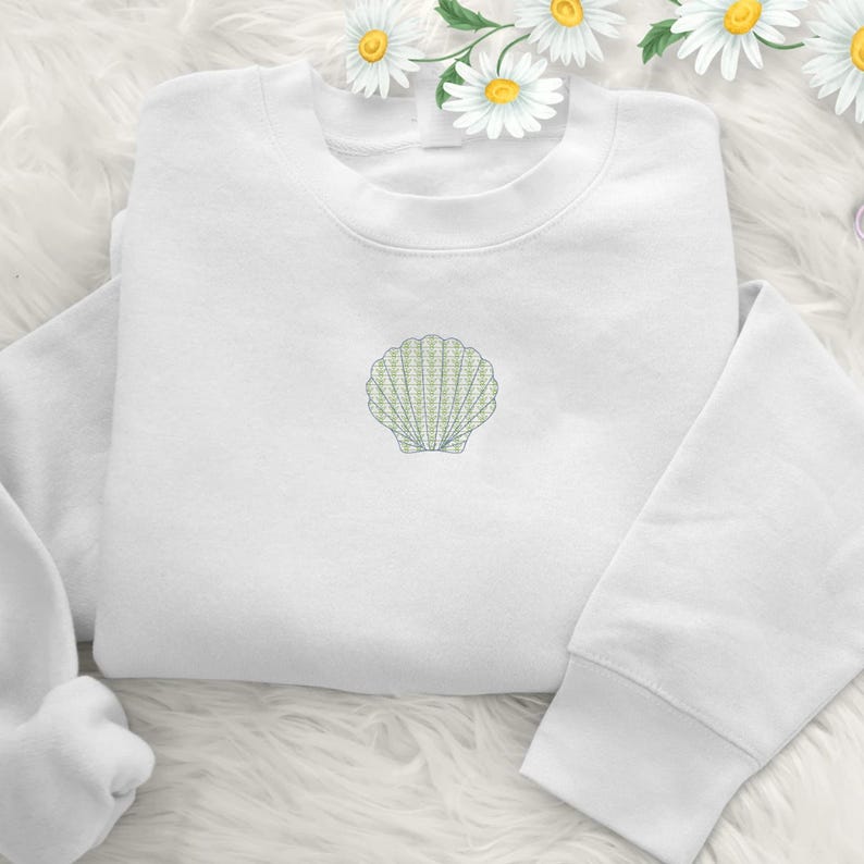 May include: A white sweatshirt with a light green embroidered seashell design on the front. The long-sleeved top is folded and placed on a white, fluffy surface. The seashell is a detailed, stylized illustration.
