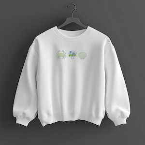 May include: White sweatshirt with embroidered designs of a crab, blue flowers, and a seashell. The sweatshirt has a crew neck and long sleeves. The embroidered designs are in shades of green and blue.