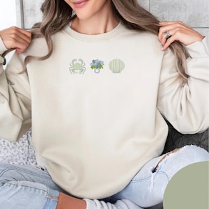May include: Cream-colored sweatshirt featuring embroidered designs of a crab, a bouquet of flowers, and a seashell. The embroidery uses green and blue thread. The sweatshirt is a casual, comfortable style.