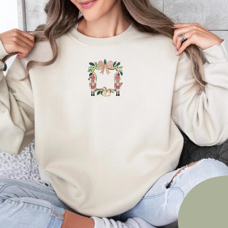 Puede incluir: Sudadera de color crema con un dise&ntilde;o bordado festivo. El dise&ntilde;o incluye un lazo rosa, hojas de acebo y dos figuras de soldados de juguete. La sudadera es de color neutro y tiene cuello redondo.