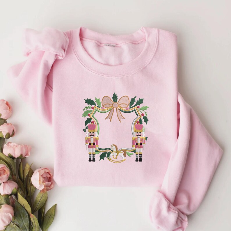 Puede incluir: Una sudadera rosa claro con un dise&ntilde;o festivo. El dise&ntilde;o incluye dos soldados de juguete, un lazo, acebo y un caballito balanc&iacute;n, enmarcado por un borde dorado. La sudadera es de un material suave y c&oacute;modo.