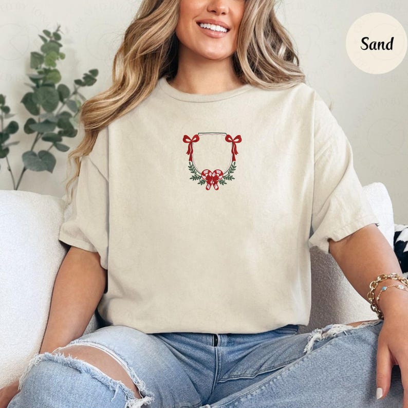 May include: Sand-colored t-shirt with a festive embroidered design. The design features a rectangular frame adorned with red bows, candy canes, and green foliage, creating a holiday wreath. The shirt is a relaxed fit.