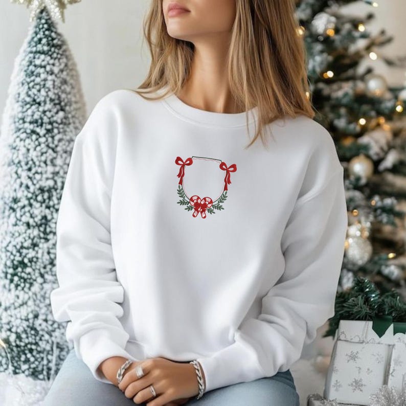 May include: White sweatshirt with a Christmas wreath design embroidered on the front. The wreath features red bows, candy canes, and green foliage. The sweatshirt is a crew neck style.
