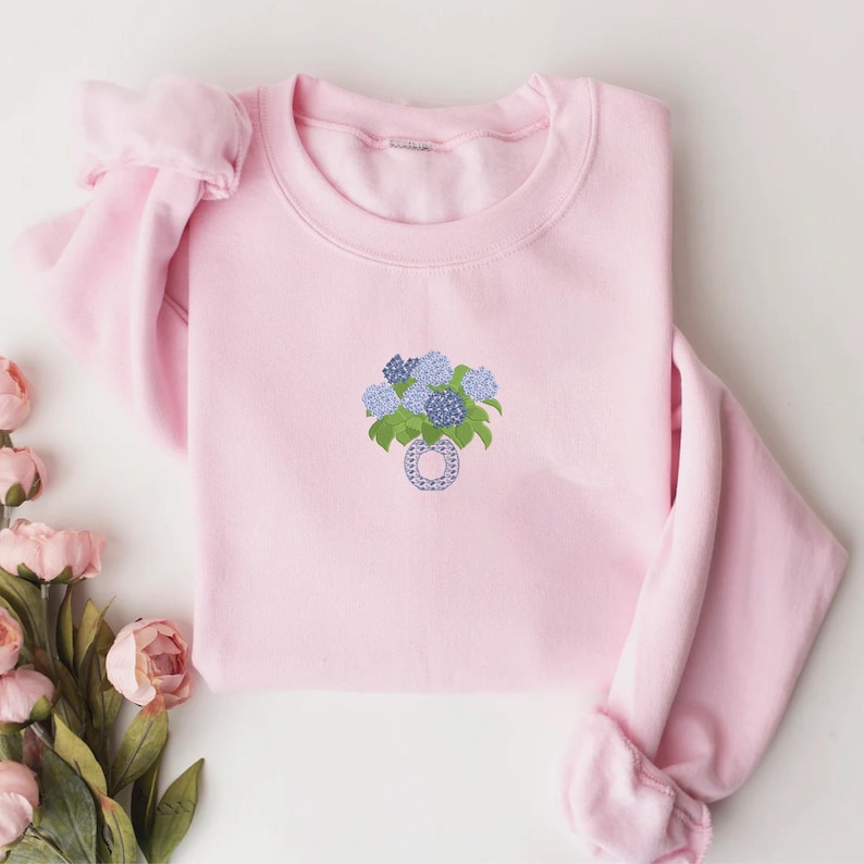 May include: A light pink sweatshirt featuring an embroidered floral design. The design includes blue and purple hydrangeas in a vase. The sweatshirt has long sleeves and a crew neck.
