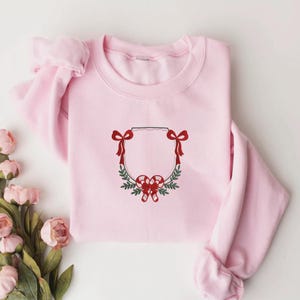 May include: A light pink sweatshirt featuring a Christmas wreath design. The wreath is composed of green leaves, red bows, and candy canes. The design is centered on the front of the sweatshirt.